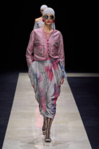 Model on runway wearing a pink patterned cropped jacket over a flowy, abstract-print skirt with wrapped waist and lace-up boots.