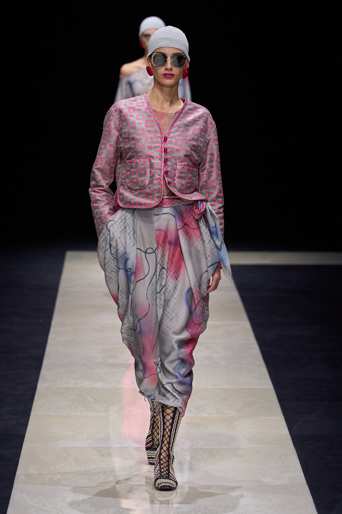 Model on runway wearing a pink patterned cropped jacket over a flowy, abstract-print skirt with wrapped waist and lace-up boots.