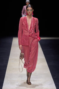 Runway model in a pink velvet suit with a deep V, pink jewelry, sunglasses, brown lace-up boots, and a small beige bag.