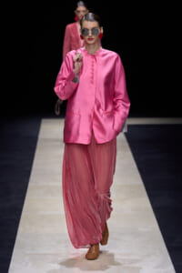 Model on a runway wearing a bright pink satin blouse with a matching sheer pink skirt and chunky red earrings, sunglasses, and braided hair.