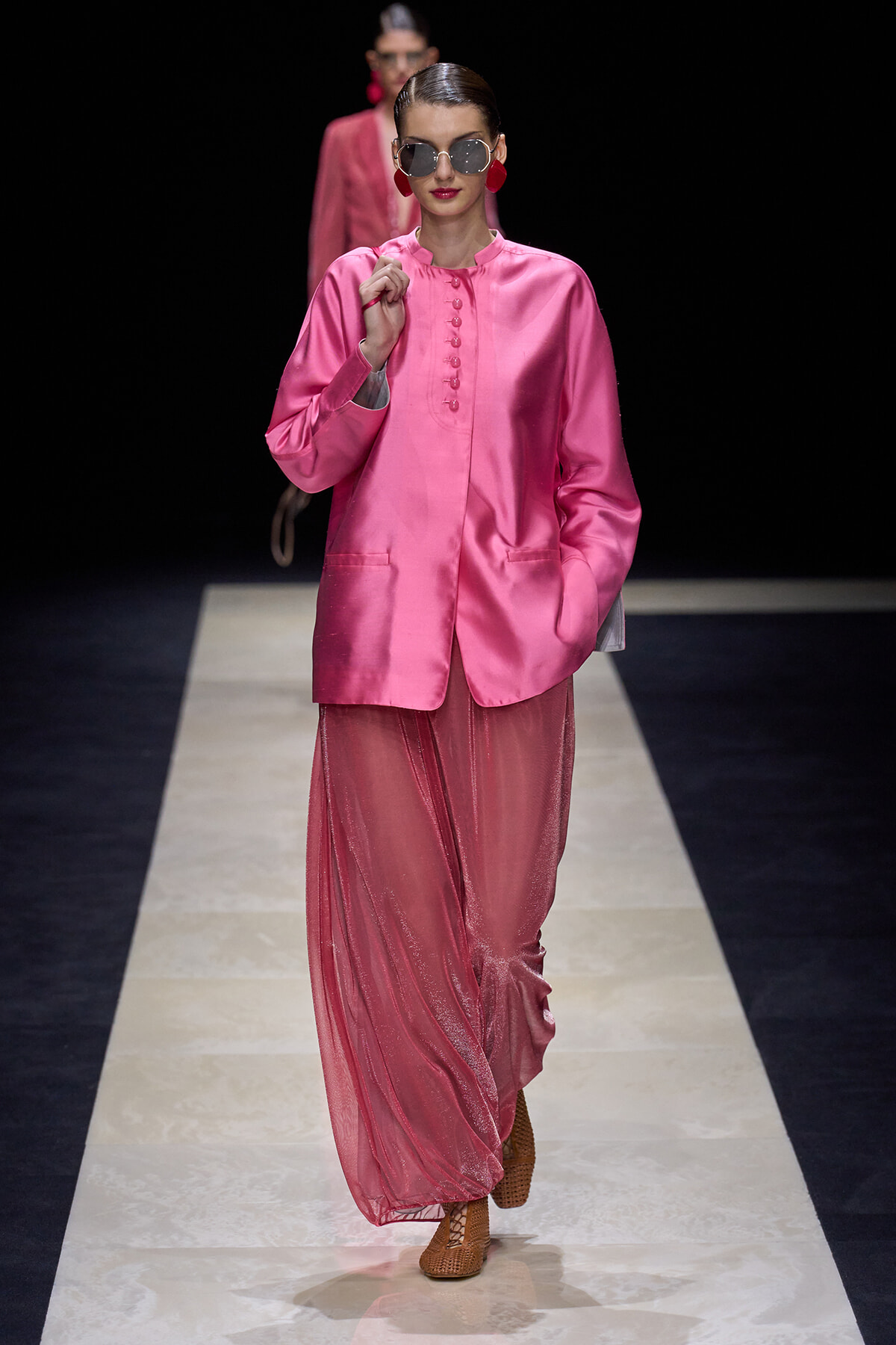 Model on a runway wearing a bright pink satin blouse with a matching sheer pink skirt and chunky red earrings, sunglasses, and braided hair.