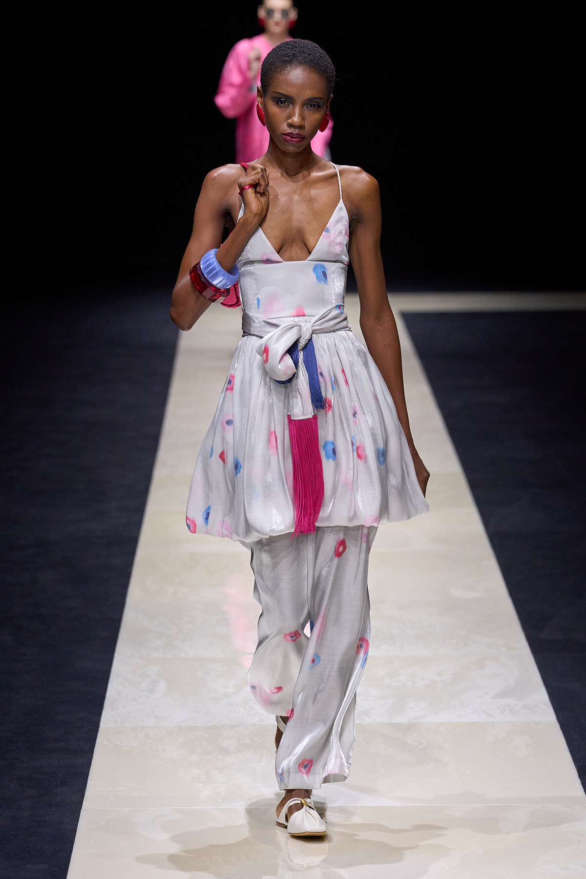 Model on a runway wearing a silver-white tie-dye dress with a waist knot, blue/pink accents, and white sandals.