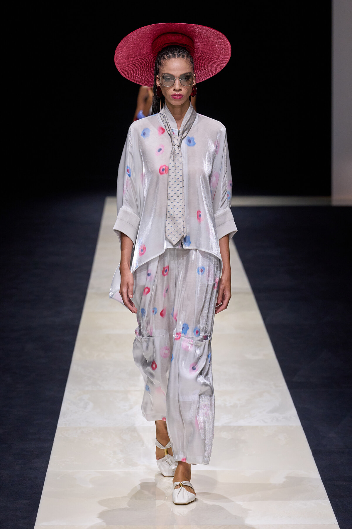 Model walks the runway in a metallic silver two-piece ensemble with scattered blue and pink spots, paired with a bright pink wide-brim hat and sunglasses.