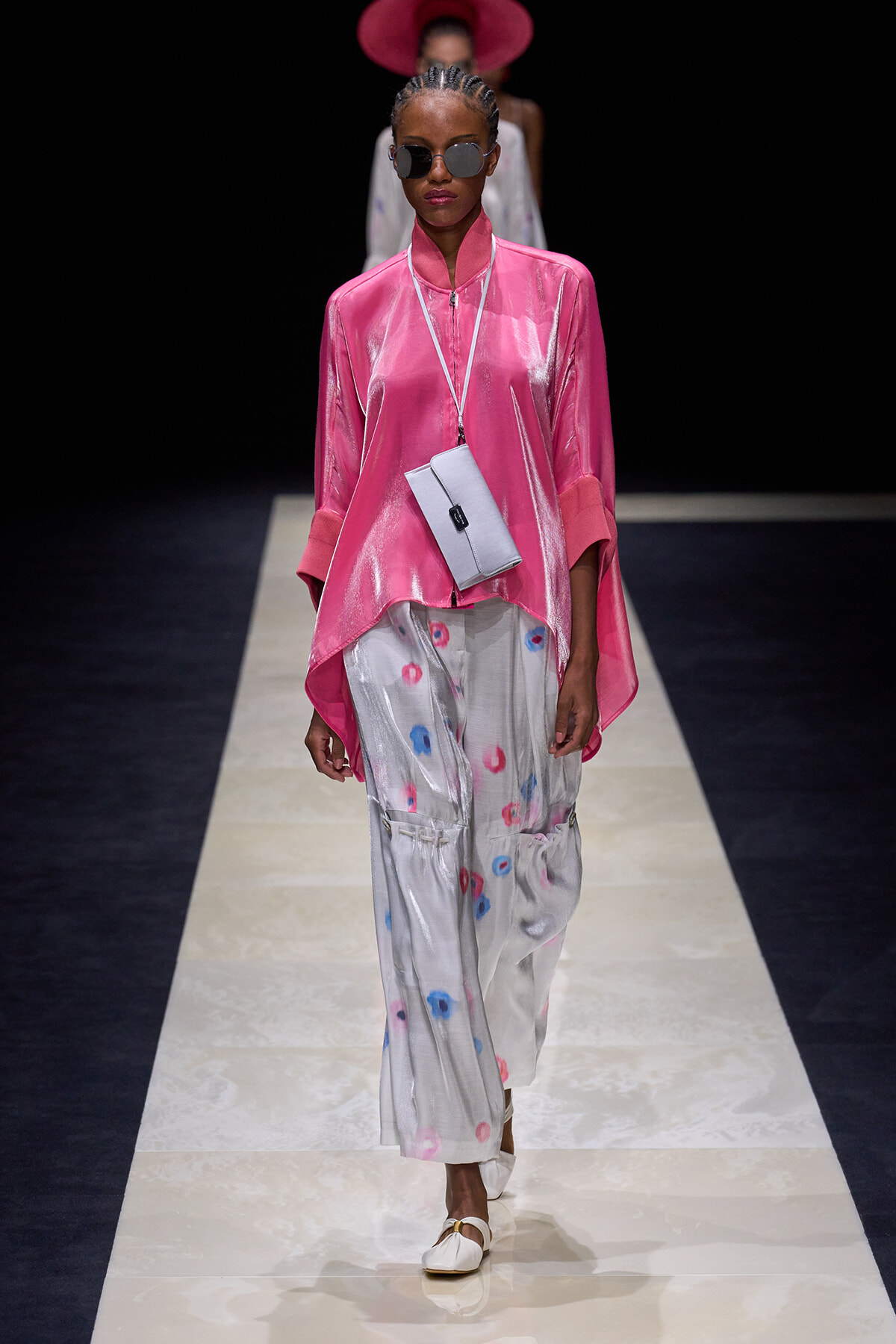 Model walking a runway in a shiny pink high-low top, silver tie-dye pants, white sandals, and a small gray crossbody bag with sunglasses and braided hair.