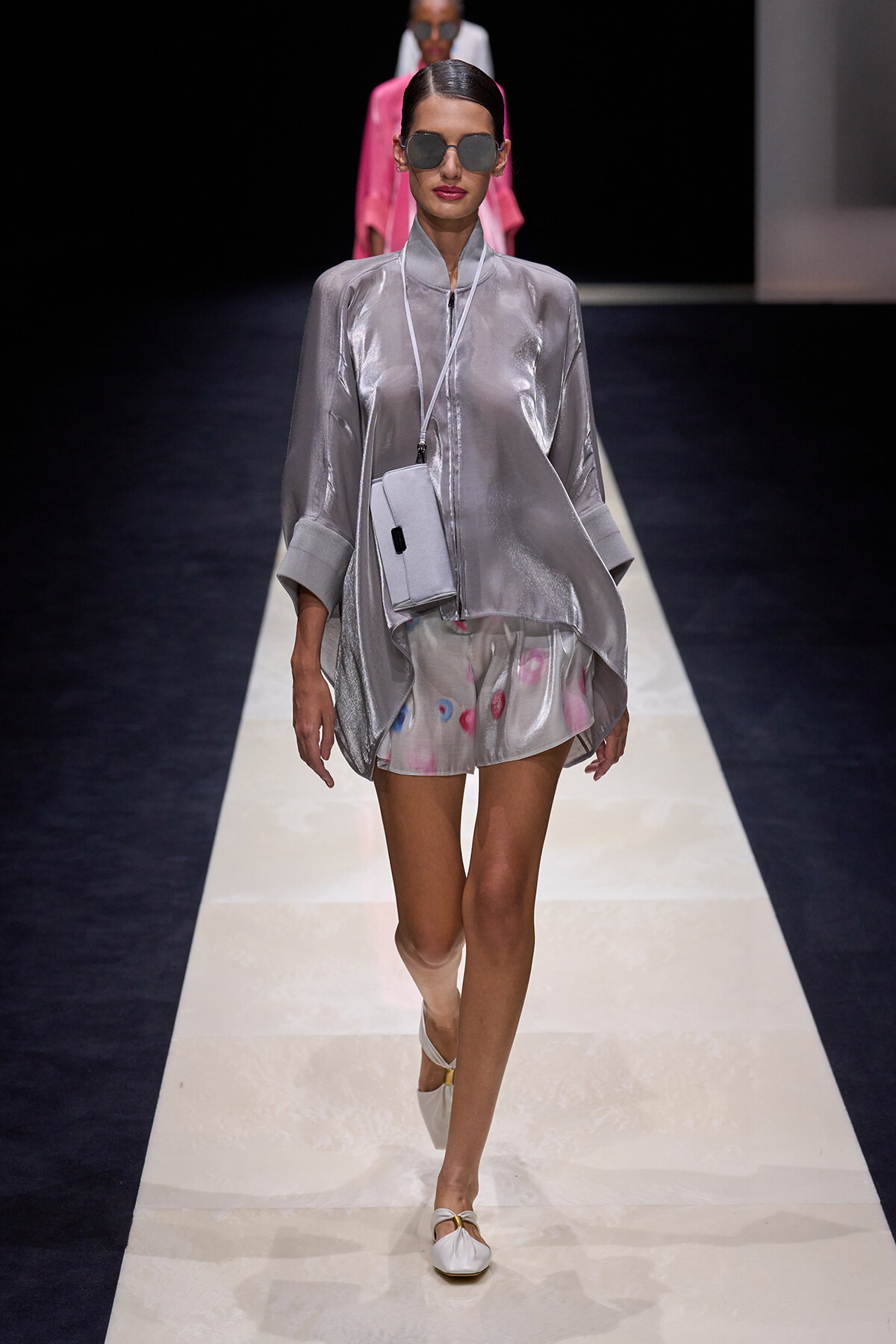Model walks a runway in a metallic silver tunic with a pale print shorts, white crossbody bag, and oversized sunglasses.