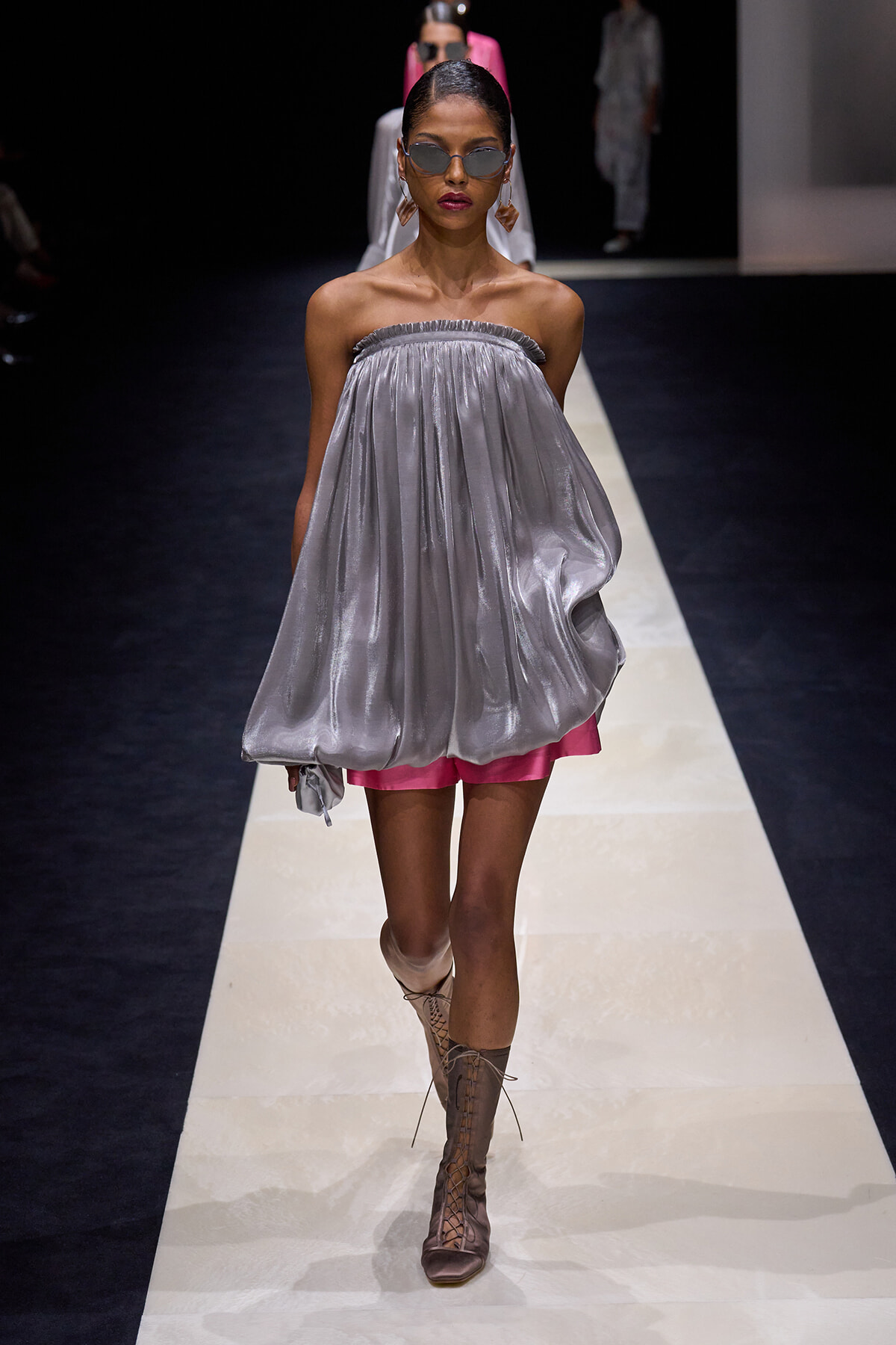 Model walks the runway in a metallic silver off-the-shoulder maxi dress layered over pink shorts, paired with beige lace-up knee-high boots and round sunglasses.