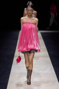Model on a runway wearing a pink strapless bubble dress, brown lace-up boots, oversized sunglasses, and a small pink handbag.