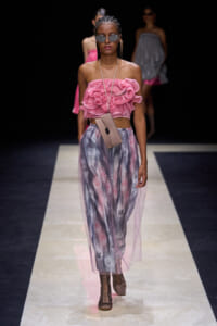 Model walks the runway in a pink ruffled strapless top with a metallic, semi-transparent skirt and a beige crossbody bag.