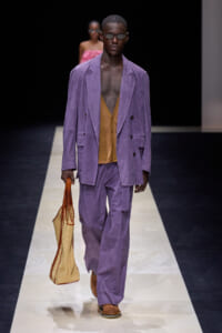 Male model walks the runway in a lavender double-breasted suit over a caramel cardigan, carrying a woven tote and wearing sunglasses.