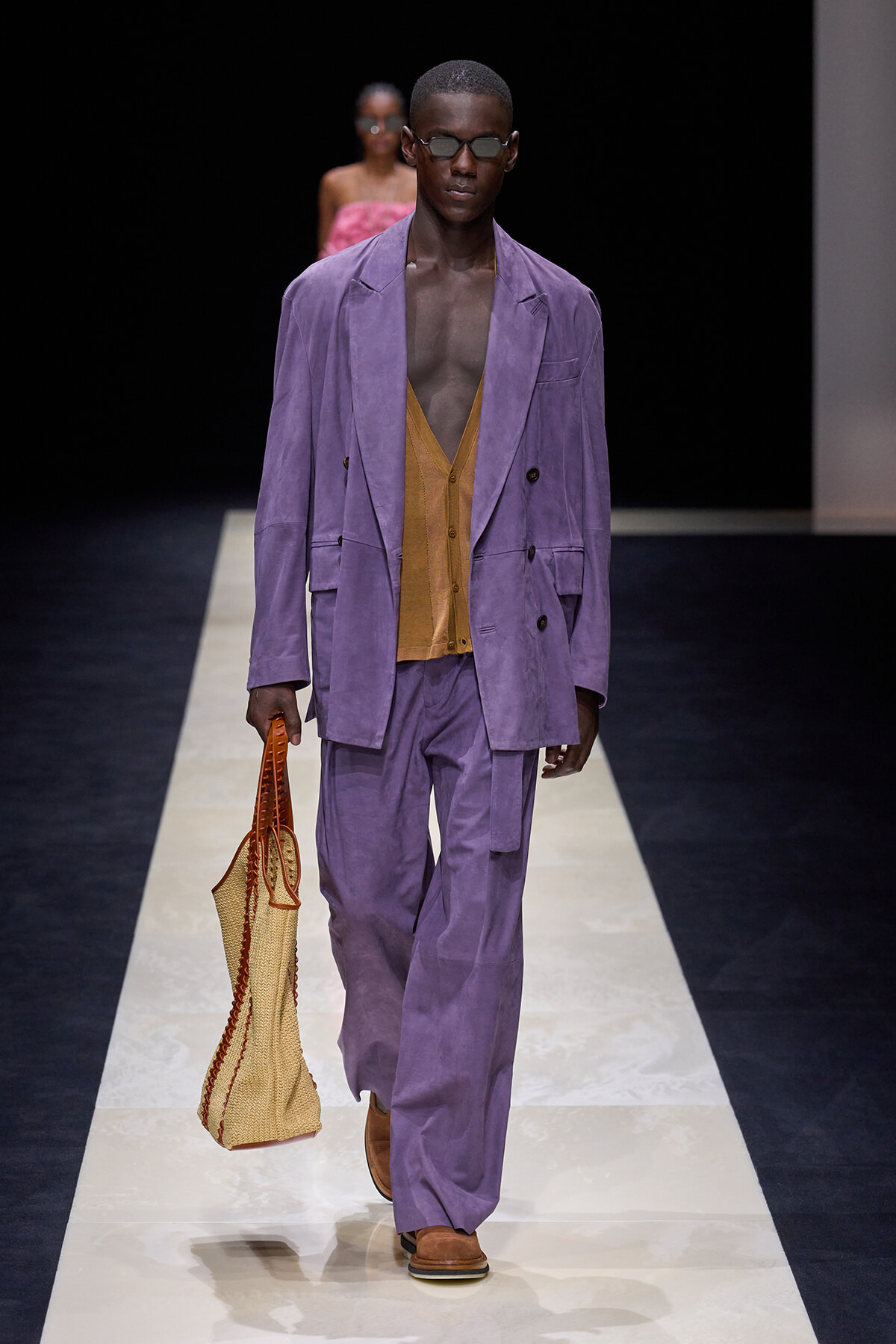 Male model walks the runway in a lavender double-breasted suit over a caramel cardigan, carrying a woven tote and wearing sunglasses.