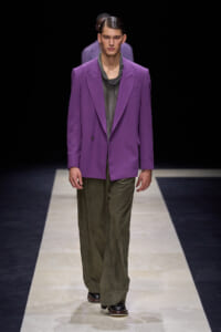 Male model walking a fashion runway in a purple blazer over a gray draped top and olive wide-leg pants on a light runway.