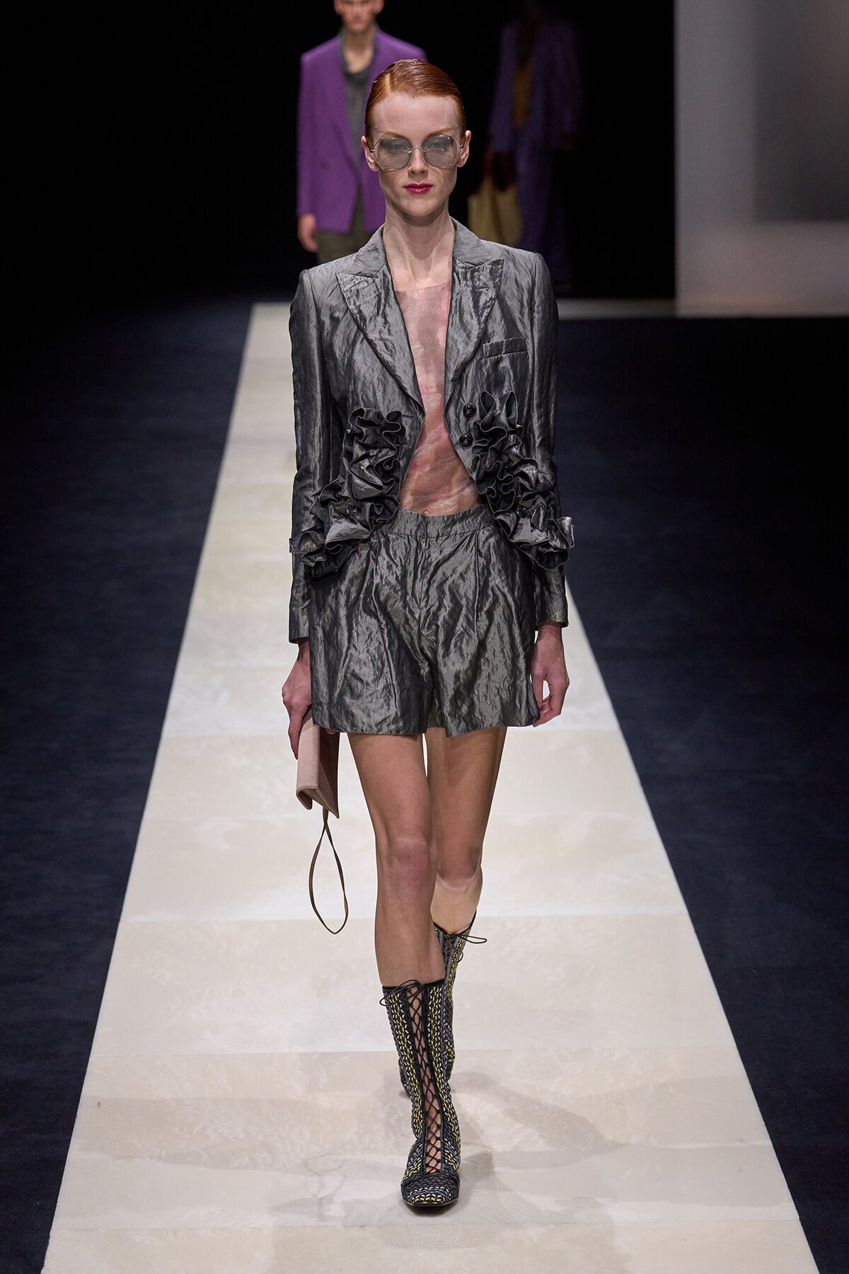 Model walking a metallic silver blazer and matching shorts with ruffled accents on the sides, sunglasses, and boots on a fashion runway.