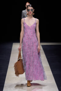 Model walking a runway in a pink lace maxi dress, tan ankle boots, oversized sunglasses, and hoop earrings, carrying a brown fringe tote.