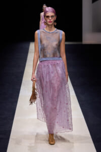 Model walking a runway in a sheer silver-lilac sleeveless top with a light purple banded waist, a long pink-lavender floral-embroidered skirt, tan ankle boots, pink headscarf, and sunglasses, carrying a small woven handbag.