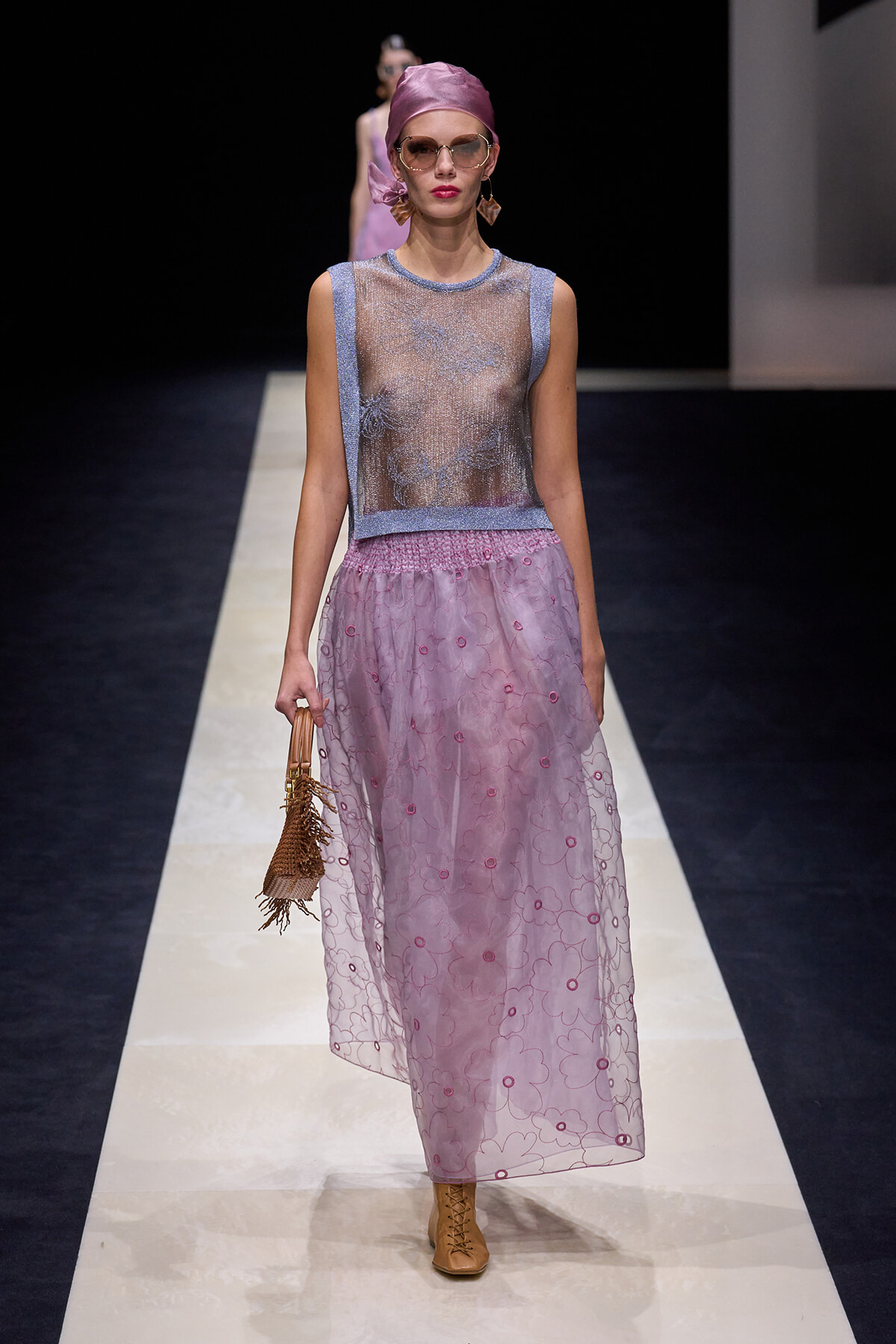 Model walking a runway in a sheer silver-lilac sleeveless top with a light purple banded waist, a long pink-lavender floral-embroidered skirt, tan ankle boots, pink headscarf, and sunglasses, carrying a small woven handbag.