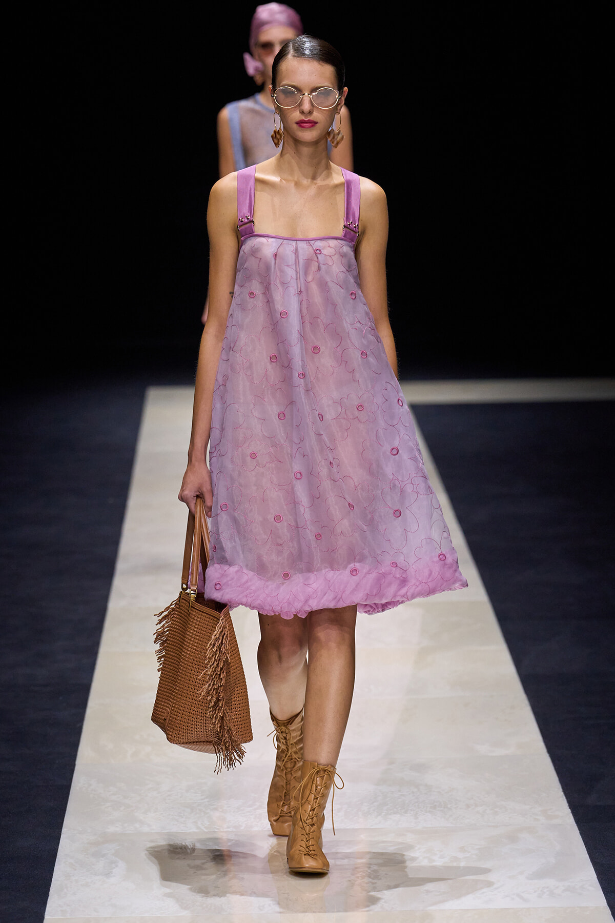Model walks the runway in a lavender pink, knee‑length sheer dress with floral embroidery and pink straps, paired with tan lace‑up boots and a brown woven tote.