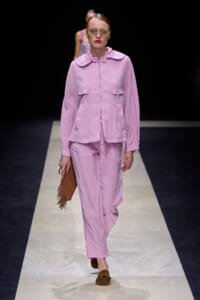 Runway model in a pastel pink hooded jacket and matching wide-leg pants, wearing round glasses and carrying a brown fringed clutch.