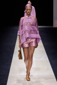 Model walks the runway in a lavender layered sheer top with matching shorts, headscarf, sunglasses, tan sandals, and a small brown handbag.