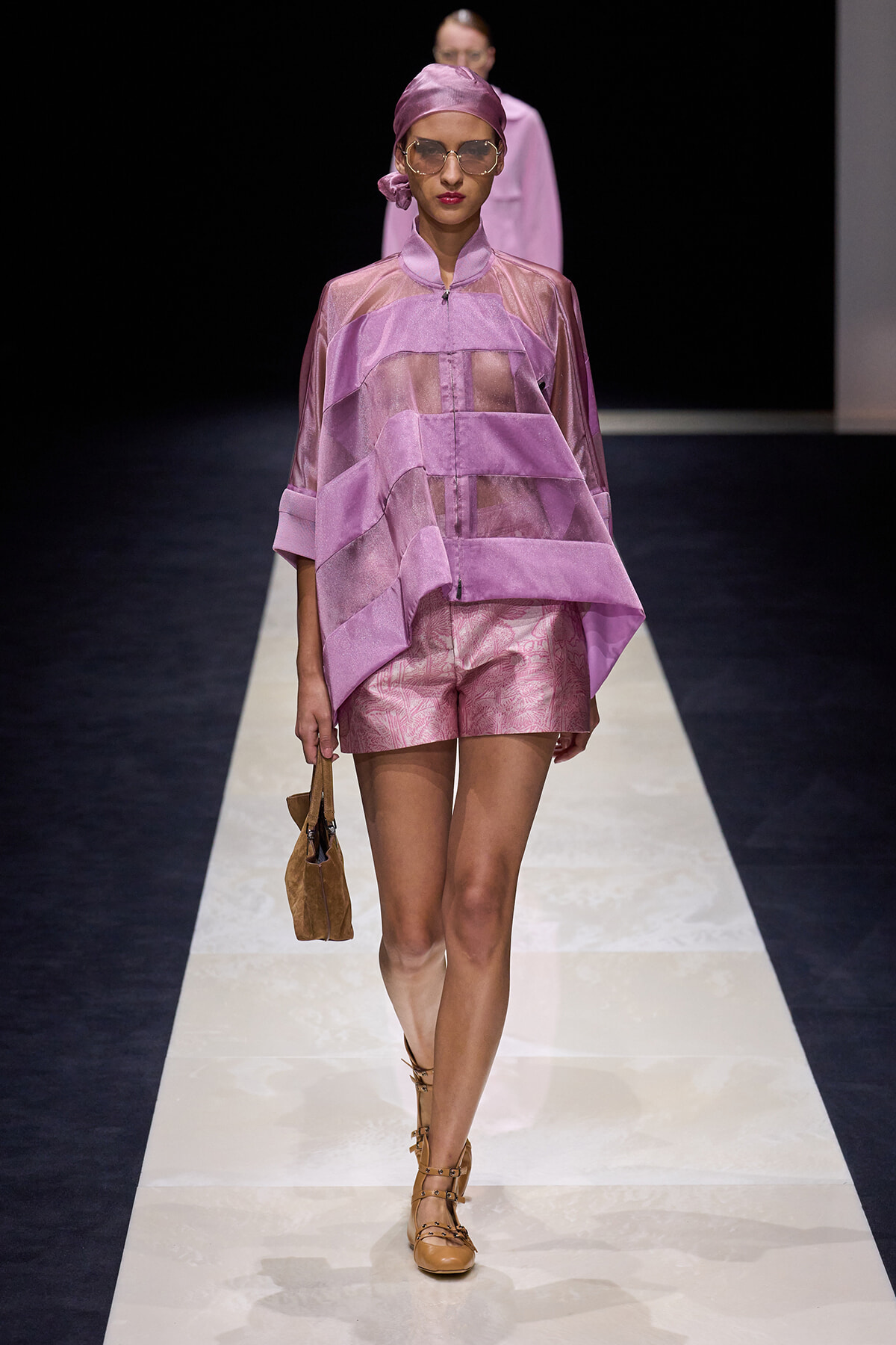 Model walks the runway in a lavender layered sheer top with matching shorts, headscarf, sunglasses, tan sandals, and a small brown handbag.