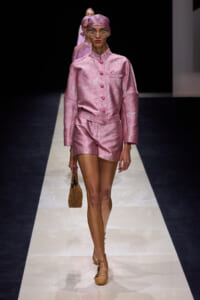 Model walking a pink satin coordinated suit (long-sleeve top with matching shorts) on a runway, wearing a pink headscarf/cap and round glasses, carrying a tan handbag.