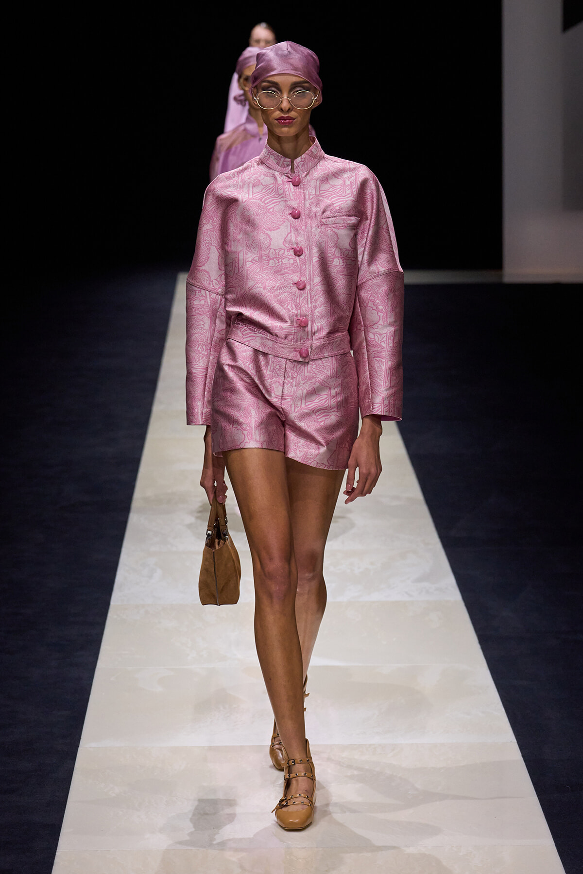 Model walking a pink satin coordinated suit (long-sleeve top with matching shorts) on a runway, wearing a pink headscarf/cap and round glasses, carrying a tan handbag.