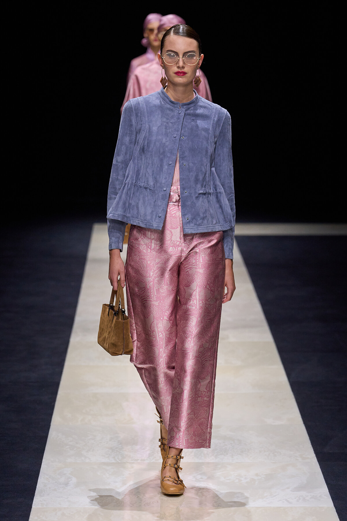 Model walking a runway in a blue suede jacket and pink brocade-style pants, carrying a brown handbag and wearing tan sandals and glasses.