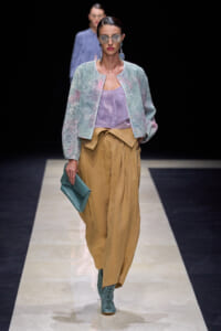 Model on a runway wearing a pastel jacket over a lavender top and tan wide-leg pants, teal lace-up boots, teal clutch, and oversized sunglasses.