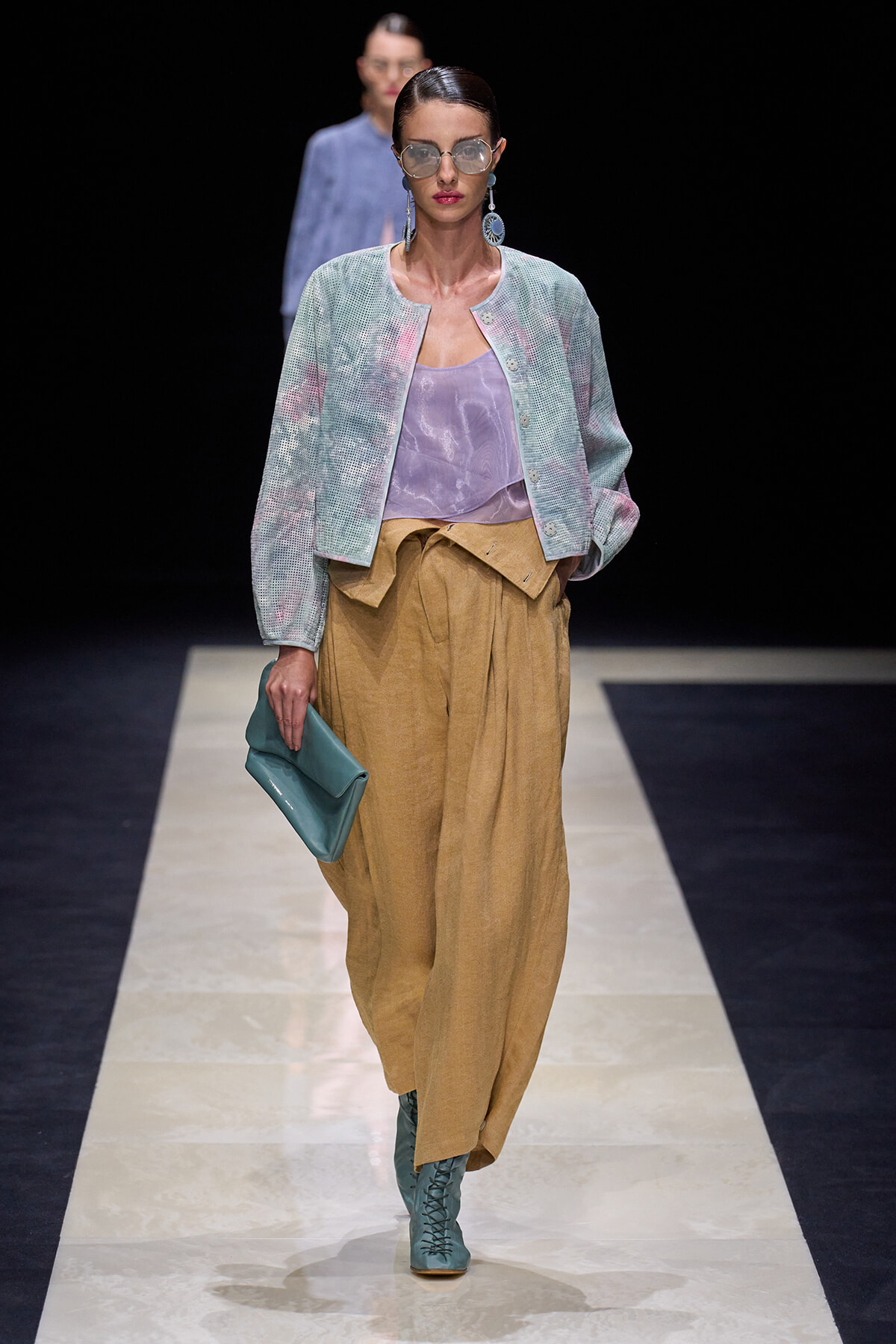 Model on a runway wearing a pastel jacket over a lavender top and tan wide-leg pants, teal lace-up boots, teal clutch, and oversized sunglasses.