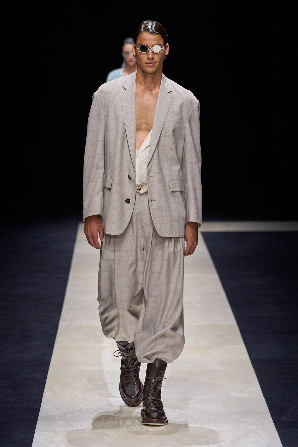 Male model in a light gray oversized suit with an open white shirt, round sunglasses, and dark boots on a runway.