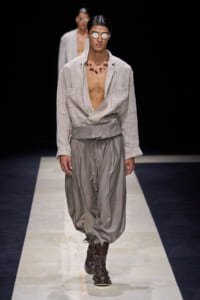 Model on a runway wearing a striped, open-front shirt with deep neckline, and oversized gray harem pants paired with brown lace-up boots and round sunglasses plus a red bead necklace.