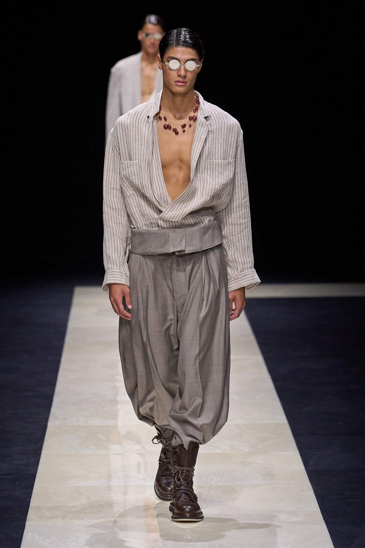 Model on a runway wearing a striped, open-front shirt with deep neckline, and oversized gray harem pants paired with brown lace-up boots and round sunglasses plus a red bead necklace.