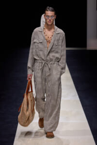 Male model walking a striped jumpsuit with a drawstring waist, sunglasses, and a beaded necklace on a fashion runway, carrying a woven tote bag.