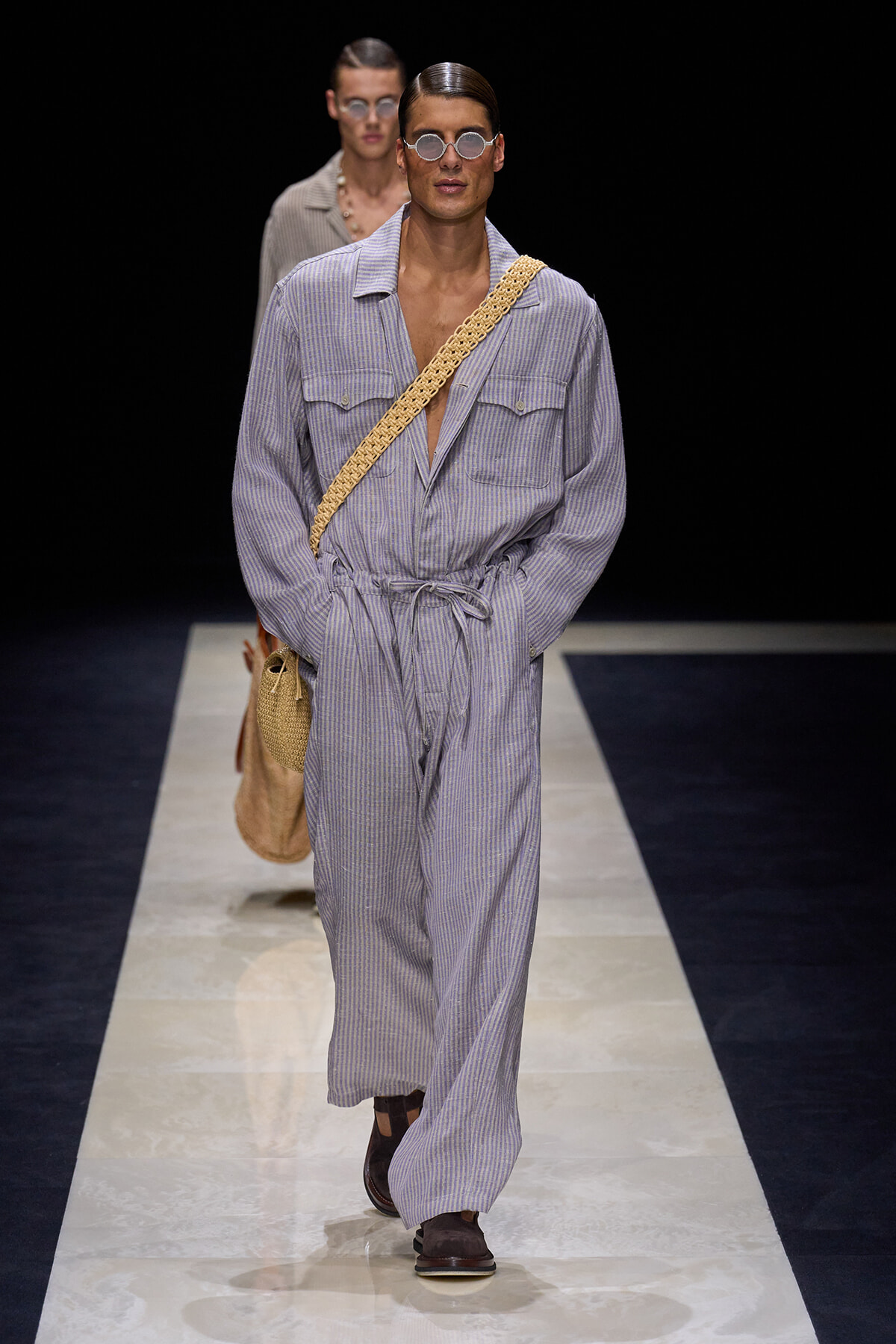 Male model walks the runway in a lavender striped jumpsuit with a tied waist, round sunglasses, and a woven crossbody bag.