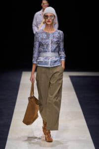 Model walking a runway in a patterned blue-gray blouse, tan wide-leg pants, tan sandals, and a brown tote bag; wearing a light knit cap and sunglasses.