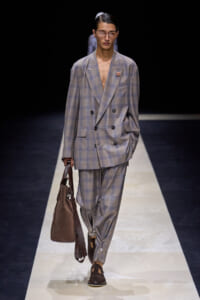 Male model on a fashion runway wearing an oversized gray plaid double-breasted suit, brown loafers, and a brown suede tote bag.