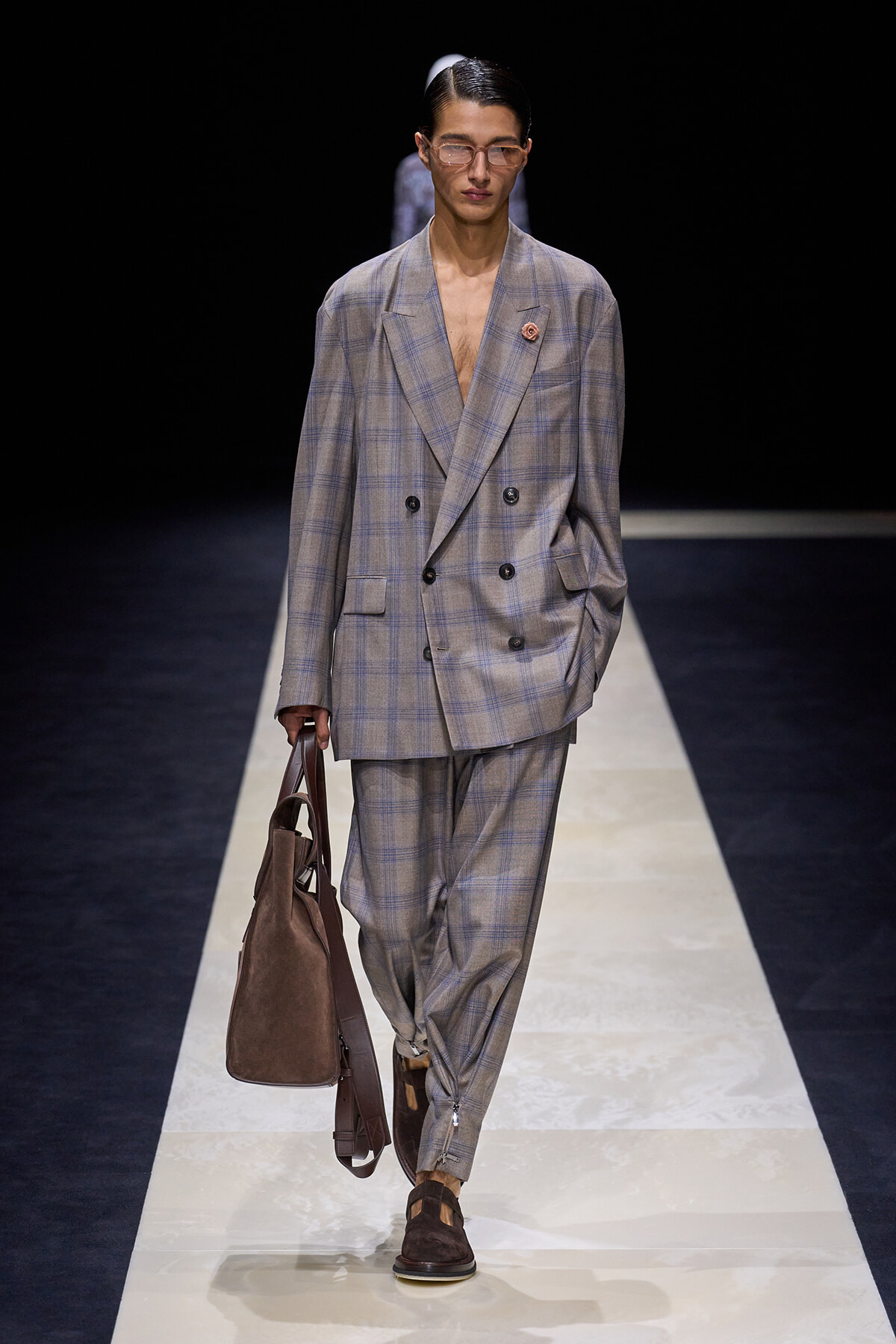 Male model on a fashion runway wearing an oversized gray plaid double-breasted suit, brown loafers, and a brown suede tote bag.