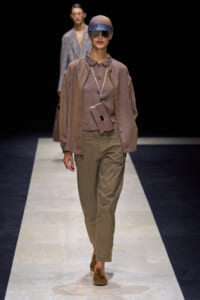 Model walks a fashion runway in a taupe oversized top, olive trousers, cap, sunglasses, and a small beige crossbody pouch.