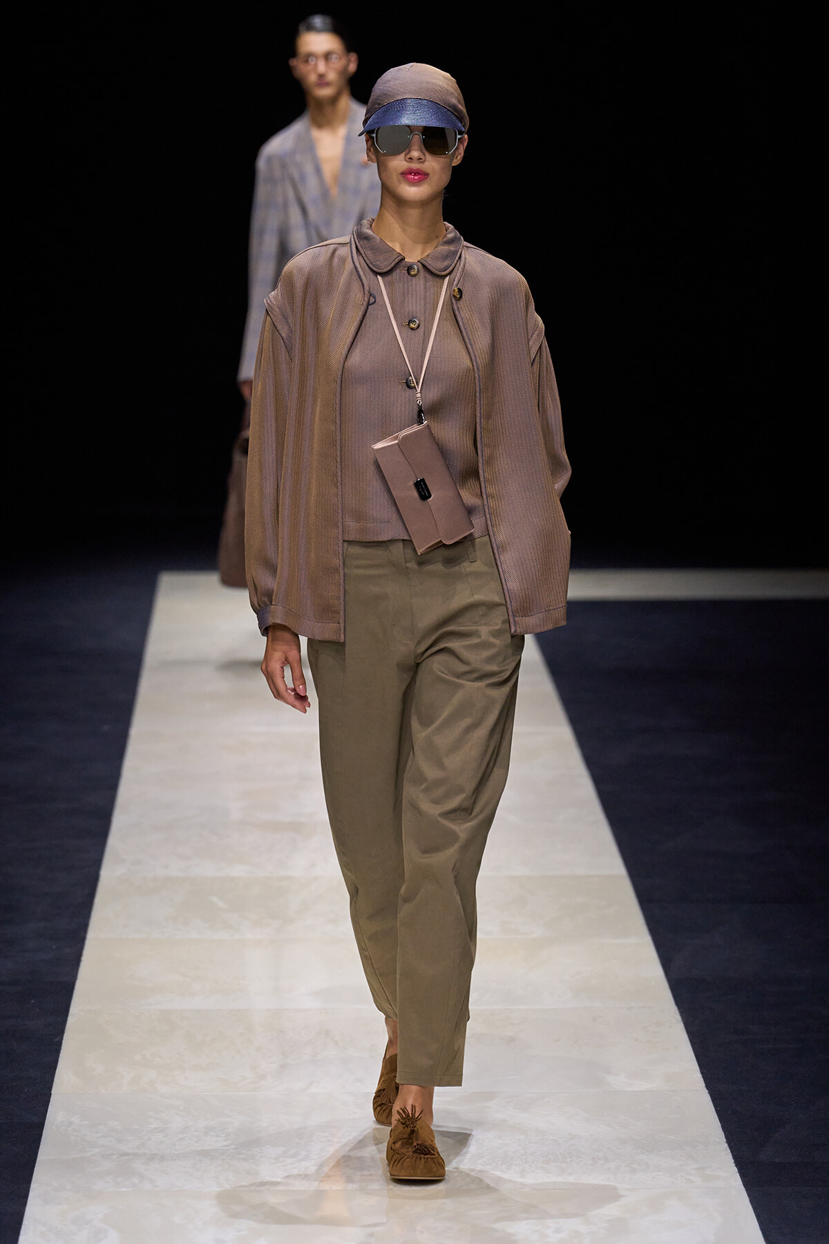 Model walks a fashion runway in a taupe oversized top, olive trousers, cap, sunglasses, and a small beige crossbody pouch.