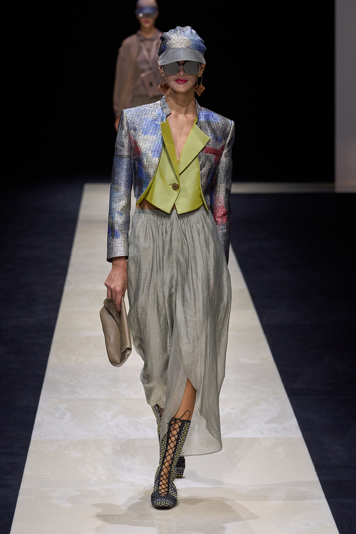 Model on a runway wearing a metallic silver blazer with a lime-green cropped vest, light gray draped skirt, cap, oversized sunglasses, and lace-up boots with a beige clutch.