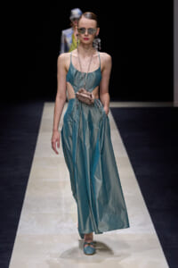 Model walking a runway in a teal cut-out satin maxi dress with a small beige pouch at the waist, sunglasses, and large circular earrings.
