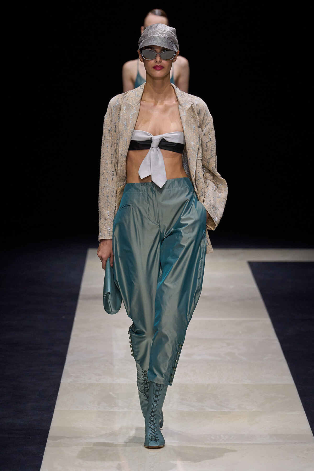 Model walking runway in beige textured blazer, gray bandeau top with knot, teal high-waisted pants, and matching lace-up boots with cap visor sunglasses and cap; dark backdrop.