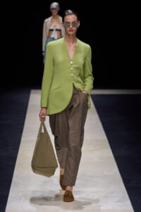 Model walking runway in light-green blazer and taupe trousers, carrying a tan tote bag; wearing sunglasses and brown loafers