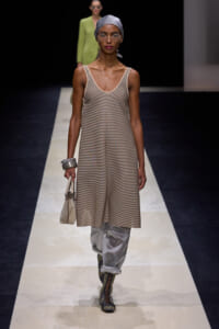 Runway model wearing a beige striped sleeveless dress over white pants, with a gray headscarf, glasses, and a small beige handbag on the runway.