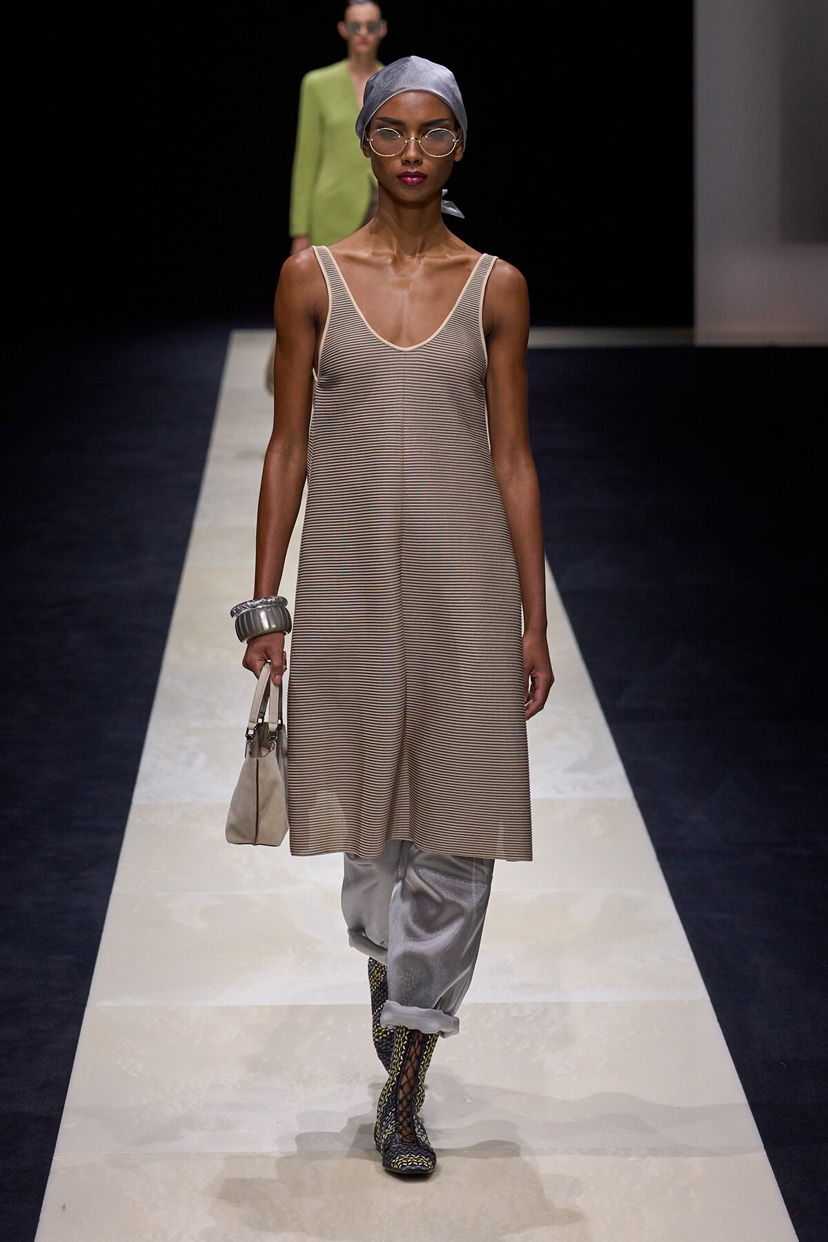 Runway model wearing a beige striped sleeveless dress over white pants, with a gray headscarf, glasses, and a small beige handbag on the runway.
