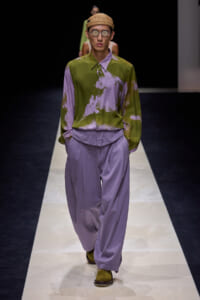 Male runway model wearing a color-blocked olive-green and lavender tie-dye shirt with lavender wide-leg pants, a straw hat, and round sunglasses as he walks the catwalk on a fashion runway.