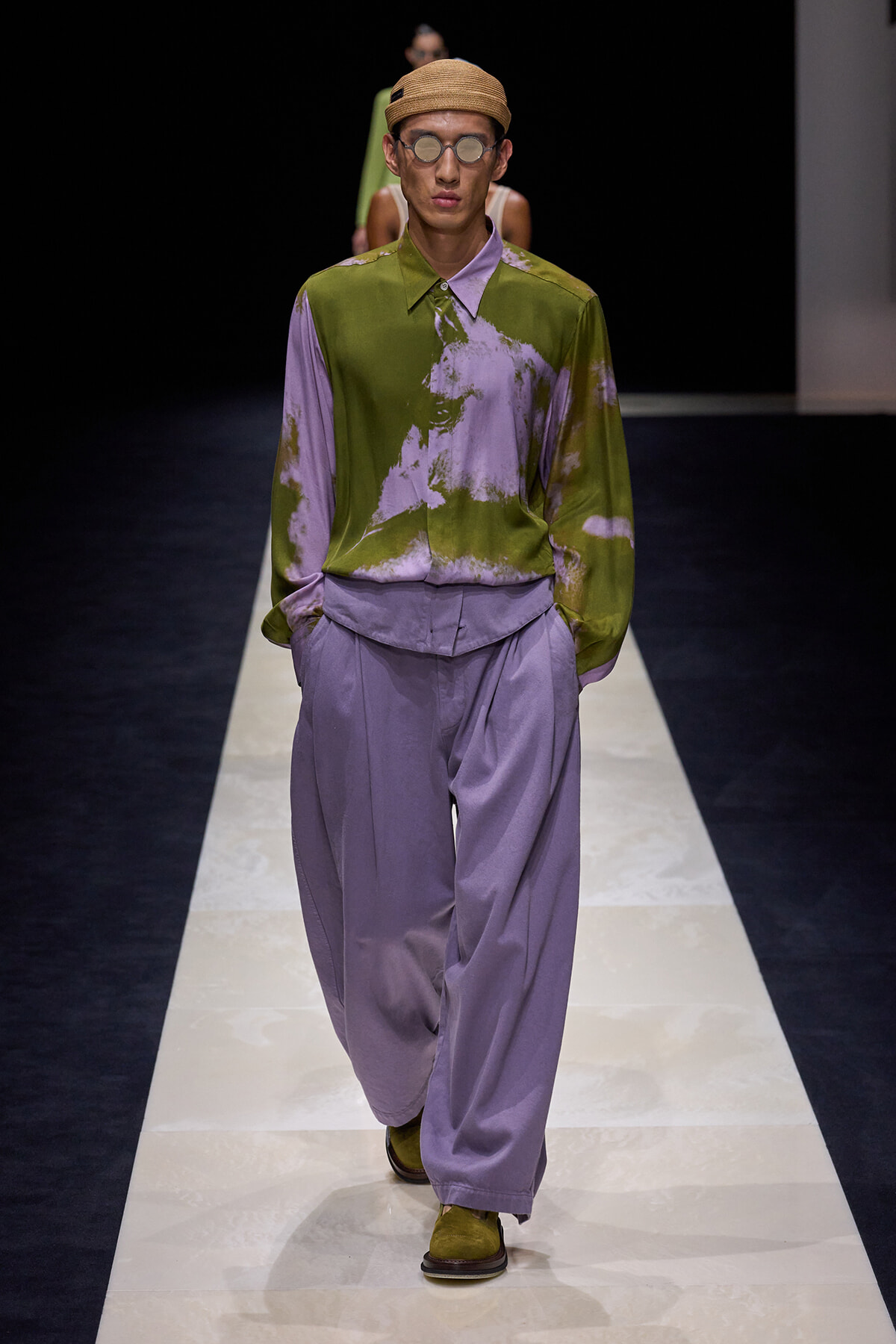 Male runway model wearing a color-blocked olive-green and lavender tie-dye shirt with lavender wide-leg pants, a straw hat, and round sunglasses as he walks the catwalk on a fashion runway.