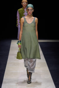 Model on a runway wearing a loose green striped sleeveless tunic over silver pants, with a green handbag and lace-up boots grouped with bracelets and a teal headscarf.