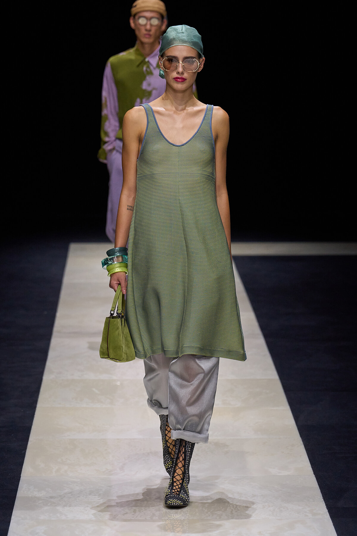 Model on a runway wearing a loose green striped sleeveless tunic over silver pants, with a green handbag and lace-up boots grouped with bracelets and a teal headscarf.