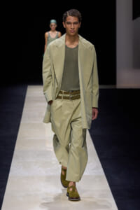 Male model walks a fashion runway in a pale green oversized blazer and matching trousers, olive mesh shirt, tan double belt, and green suede sandals.