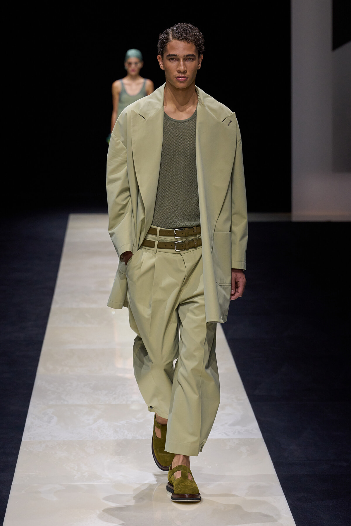 Male model walks a fashion runway in a pale green oversized blazer and matching trousers, olive mesh shirt, tan double belt, and green suede sandals.